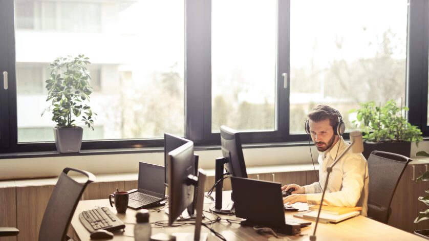 A businessman sits at a desk using multiple computers and a headset in a well-lit modern office.